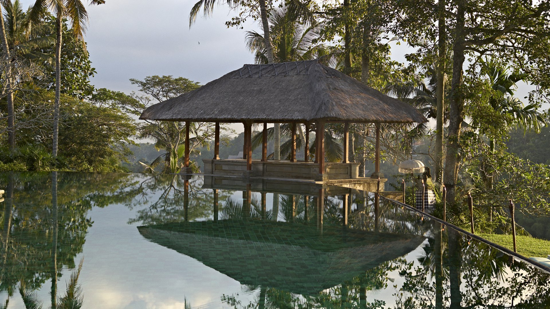 The thatched bale of Amandari reflected in its pool at dusk, among palms and tall trees